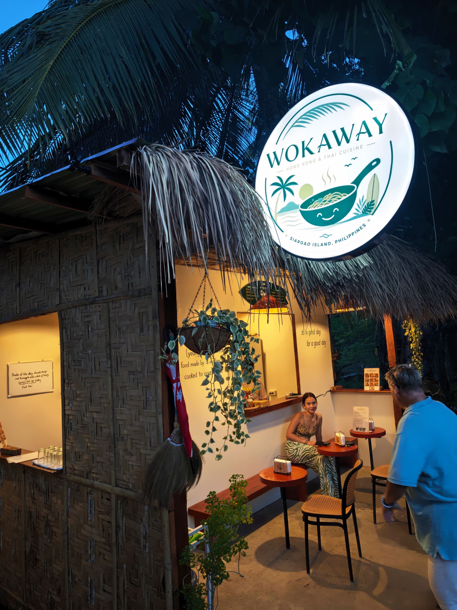 The lit Wokaway sign at dusk on Back Road, palms and a thatched roof above, guests sitting at the terracotta tables inside