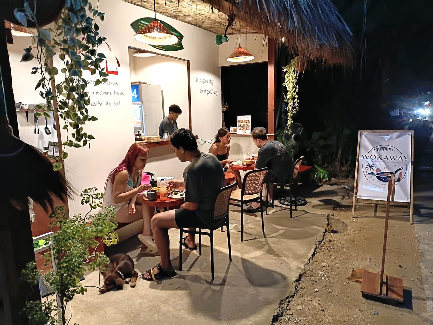 The open front of Wokaway at night, guests eating at the small red tables under woven lampshades, the Wokaway banner out by the road