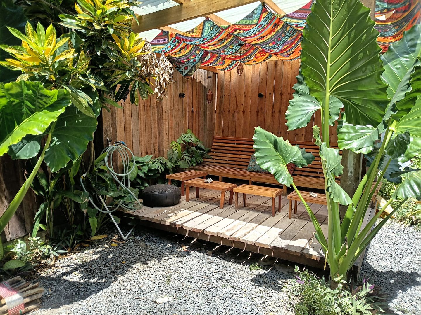 The garden in daylight: patterned bunting strung over a pergola above a timber deck and bench, framed by large elephant ear leaves