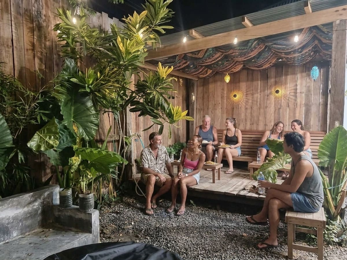 The garden behind the kitchen after dark, guests eating and talking on the timber deck and benches under strings of warm bulbs, dense tropical planting around them