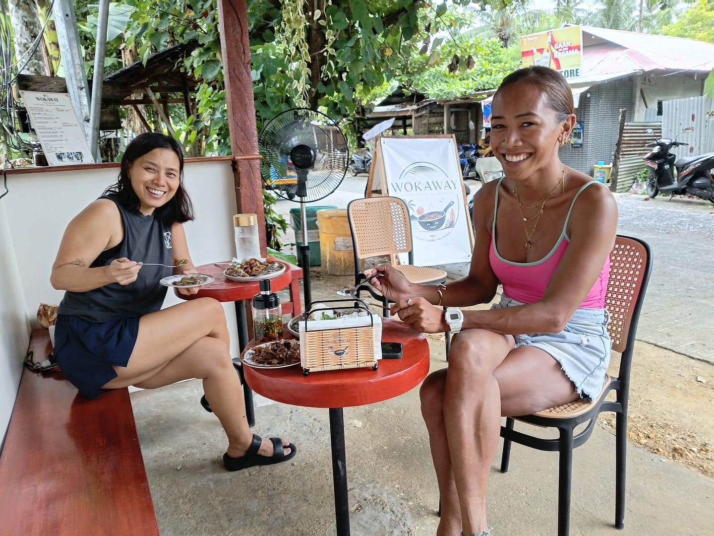 Two guests eating at the red tables under the roadside canopy, the Wokaway banner and the printed menu on the wall behind them