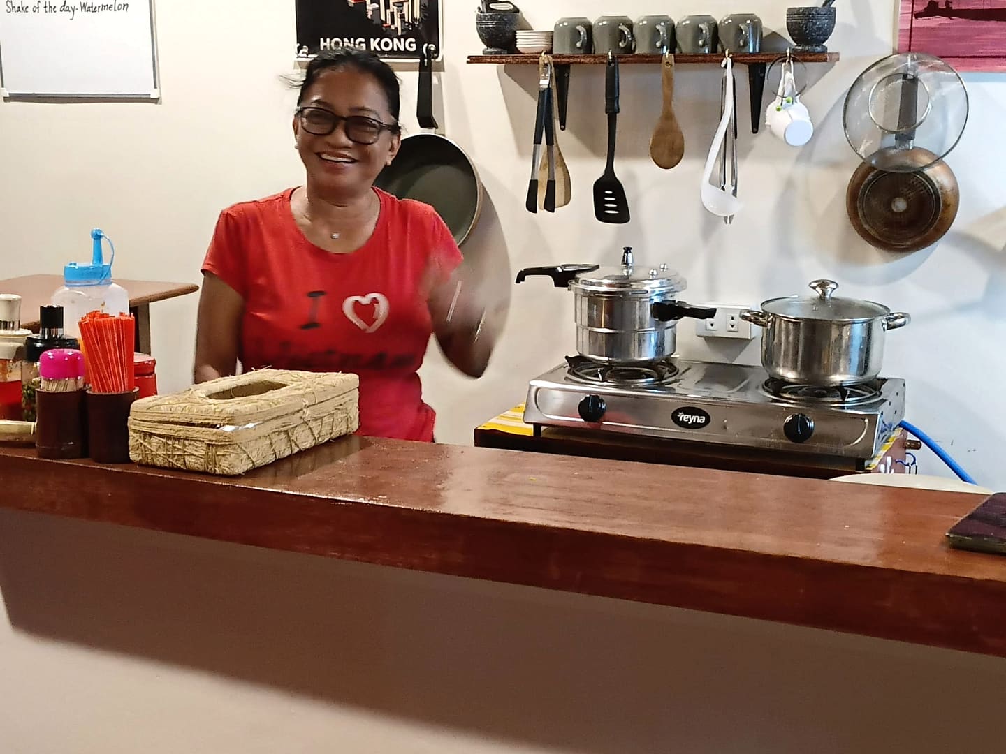 The cook at the kitchen counter with a wok lid in her hand, a Hong Kong poster and rows of hanging utensils on the wall behind