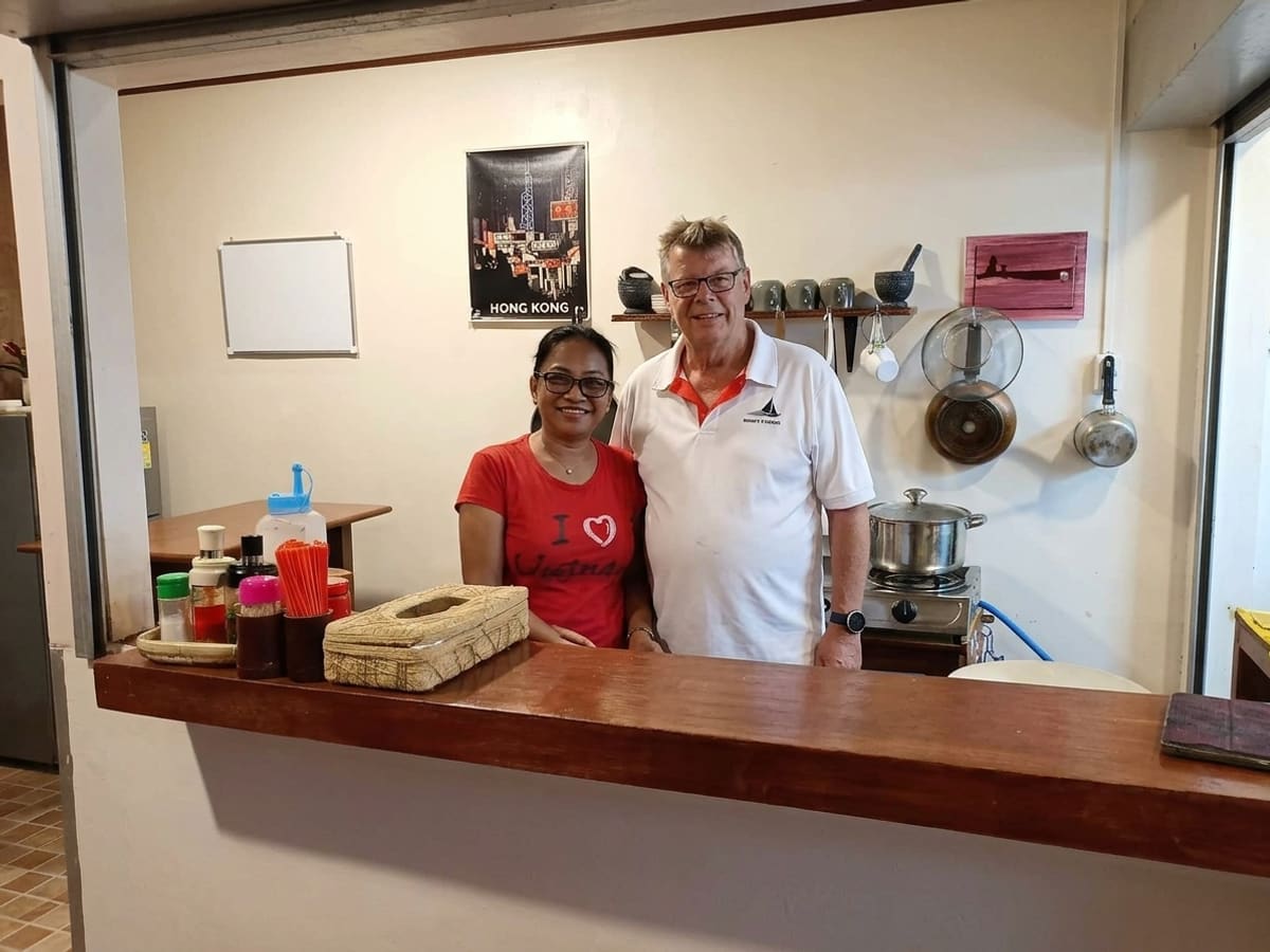 The two owners standing together behind the counter of their kitchen, the Hong Kong poster and the shelf of cups behind them