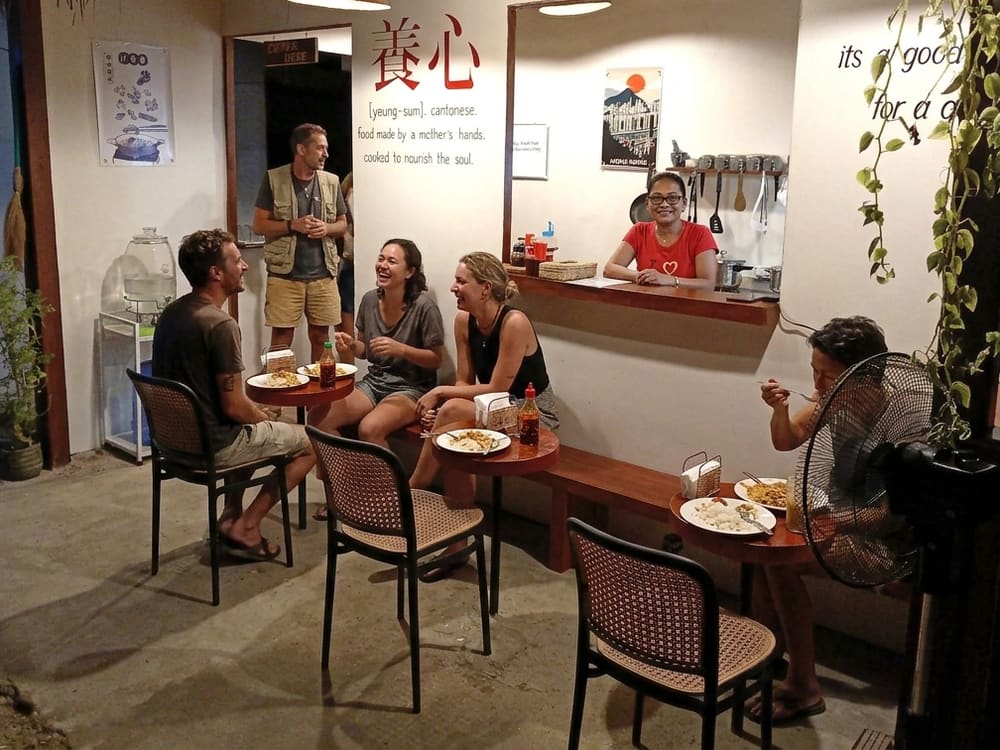 Guests eating and laughing at the counter tables under the painted 養心 wall, the cook smiling from the serving hatch behind them